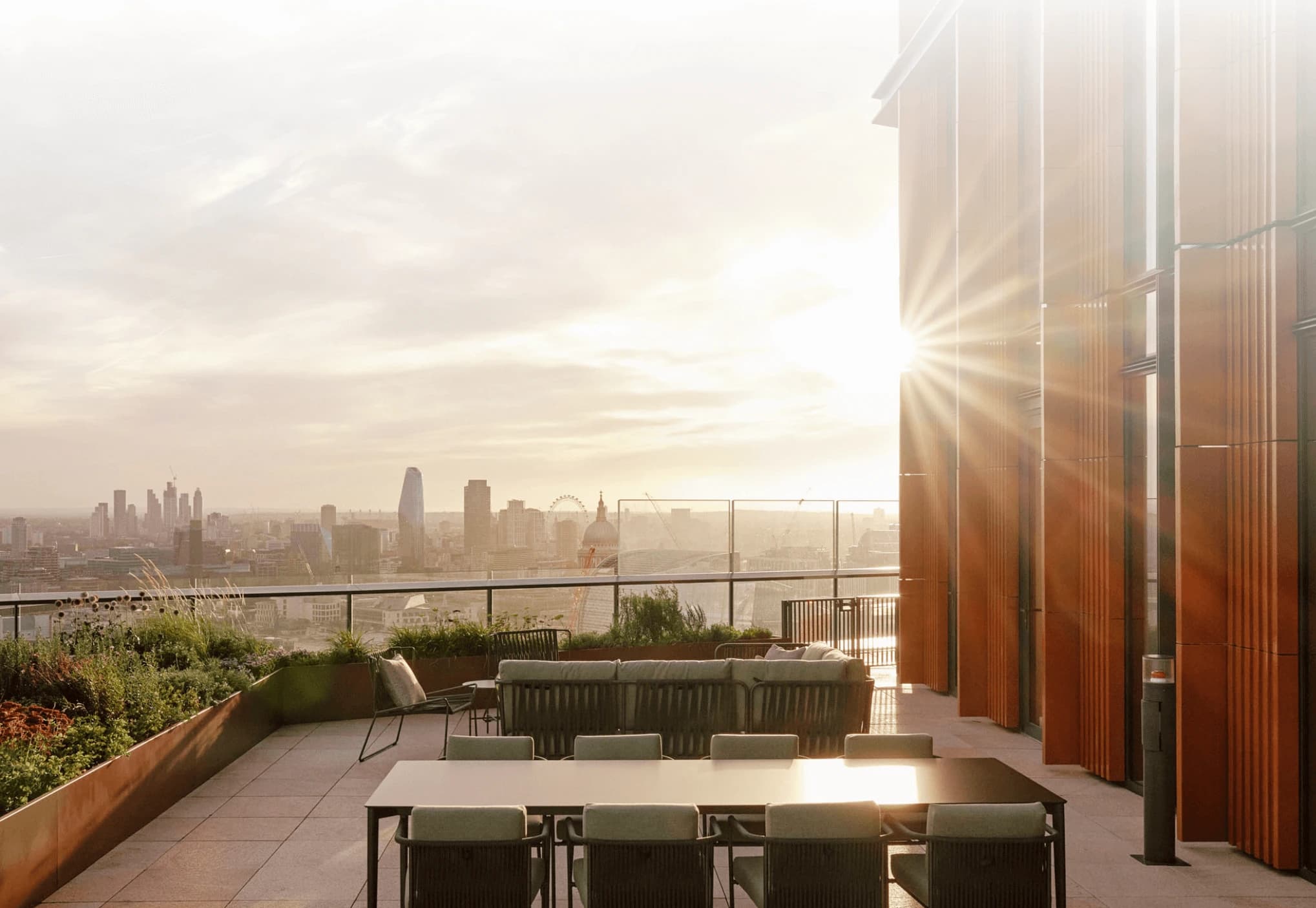 Rooftop terrace at sunset with outdoor dining table and lounge seating overlooking the London skyline, sunlight flaring beside a modern building facade.